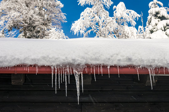 View of icicles hanging from a snowy rooftop of a wooden mountain cottage, hut or house with trees covered in snow and clear blue sky in the background, beautiful winter or Christmas scene