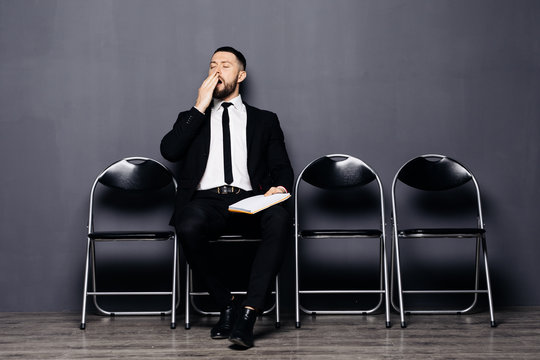 Serious bearded young man in glasses and formal wear is yawing while sitting in the hall on chair