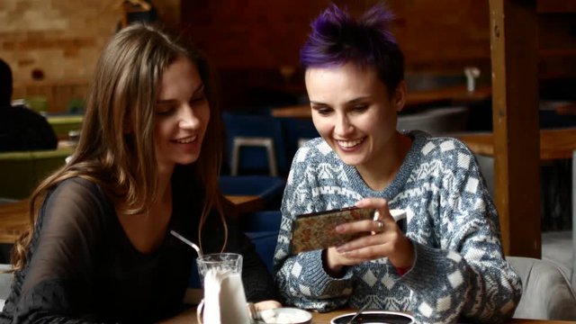 Two friends in a cafe watch video on the smartphone and laugh. Women drink coffee in the coffee house