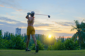 woman golf player in action of end downswing of wood driver, after hit the golf ball away from tee off to the fairway ahead, sunset scenery in background