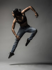Beautiful Biracial Asian Black Dancer wearing blue jeans and baseball cap does a creative urban dance move against a grey backdrop in studio with dramatic lighting