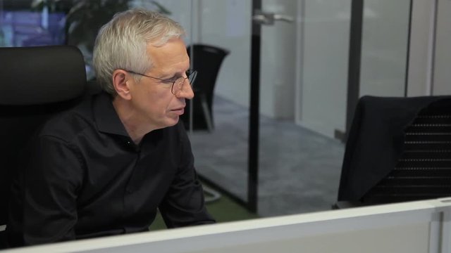 Senior chief working while sitting at computer in modern office. Man in glasses is in leather armchair and looks attentively at PC screen, reading important data