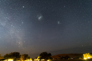 Fototapeta premium The starry sky and the majestic Magellanic Clouds, outstandingly bright, captured in Africa. Acacia trees and straw hut in the foreground. Adventure into the wild.