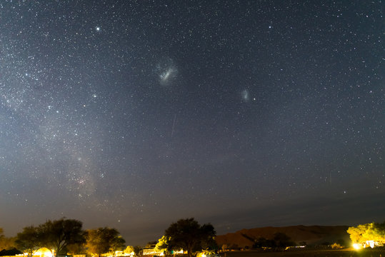 The starry sky and the majestic Magellanic Clouds, outstandingly bright, captured in Africa. Acacia trees and straw hut in the foreground. Adventure into the wild.