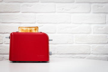 Red toaster with toasted bread for breakfast inside. White table. White brick wall.