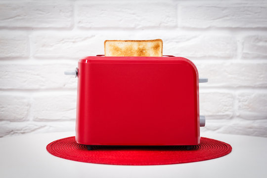 Red toaster with toasted bread for breakfast inside. Red table napkin. White table. White brick wall.