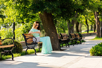 Young beautiful woman in a light green pastel long dress is sitting on a park bench and is looking at a mobile phone.