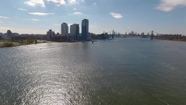 Aerial real time shot of New York City. The camera approaching to Williamsburg neighborhood above East River filming Williamsburg Bridge and Downtown Manhattan. A ferry is travelling along the river.