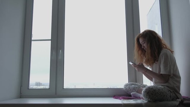 Beautiful young woman with mobile phone sitting on window sill at home. Redhead female talking on the Internet, rewritten.