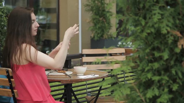 Beautiful woman doing selfie, sitting in summer cafe outdoors. Lady looks at screen of white smartphone then takes photo of herself, smiling amicably. Attractive brunette with long hair, dressed in