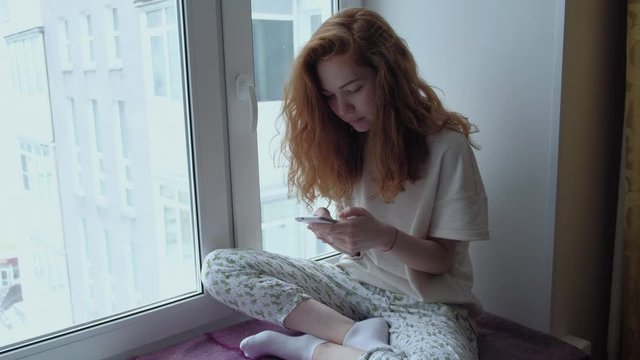 Young redhead female sitting on the windowsill with a mobile phone.Beautiful young woman with mobile phone sitting on window sill at home. Redhead female talking on the Internet, rewritten.