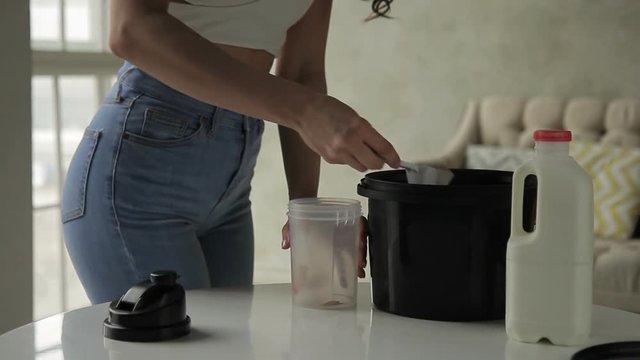 Young woman pours whey into shaker while standing in house kitchen. Female opens lid of plastic container and puts dry mixture inside from jar with spoon. Fitness lady dressed in white top with bare