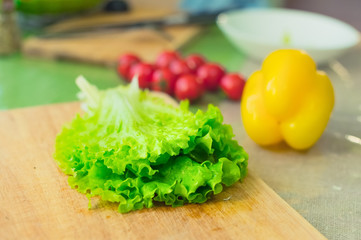 Fresh green lettuce leaves lie on a wooden cutting board next to the yellow bell pepper and red cherry tomatoes on a green table
