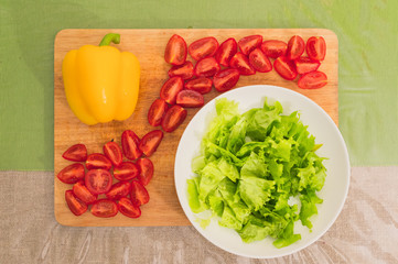 Fresh chopped green lettuce leaves lie on a wooden cutting board next to the yellow bell pepper and chopped red cherry tomatoes on a green table