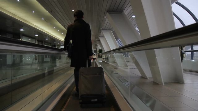 The korean man stands on travelator in the big broght corridor of modern airport. The brunette male wears expensive black suit and holds the handle of his useful luggage bag while slow moving of the