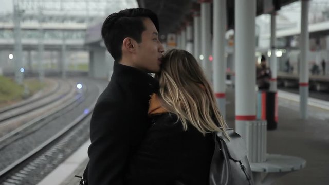 The happy multiethnic couple stands in the train station, talks and hugs enjoying their meeting. The asian man wears dark coat and his girlfriend has round silver glasses, red lipstic and orange scarf