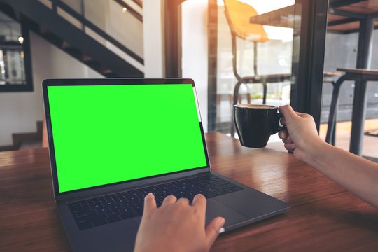 Mockup image of a hand using and touching laptop with blank green screen while drinking coffee on wooden table in cafe