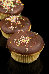 Vanilla cupcakes with chocolate cream and colorful sprinkles above black reflections background