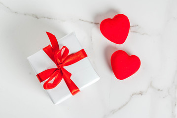 Valentine's day concept, white wrapped gift box with red ribbon and red velvet hearts, on white marble background, copy space top view