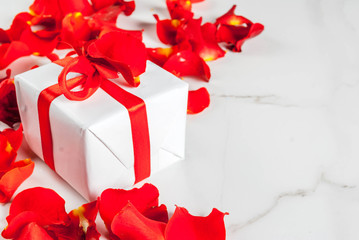 Valentine's day concept, with rose flower petals and white wrapped gift box with red ribbon, on white marble background, copy space