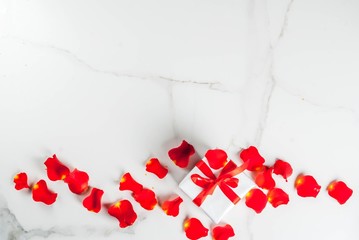 Valentine's day concept, with rose flower petals and white wrapped gift box with red ribbon, on white marble background, copy space top view