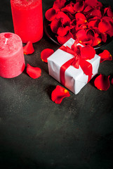 Valentine's day background with rose flower petals, white wrapped gift box with red ribbon and holiday red candle, on dark stone background, copy space