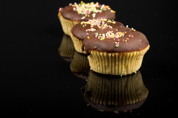 Vanilla cupcakes with chocolate cream and colorful sprinkles above black reflections background