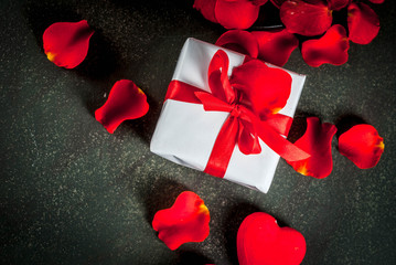 Valentine's day background with rose flower petals, white wrapped gift box with red ribbon and holiday red candle, on dark stone background, copy space top view