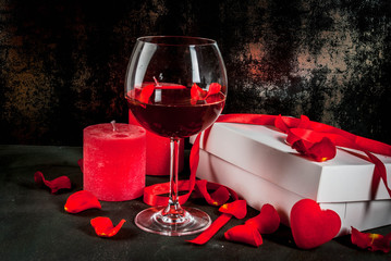 Valentine's day concept, white wrapped gift box with red ribbon, rose flower petals, red wine glass, with red candle, on dark stone background, copy space