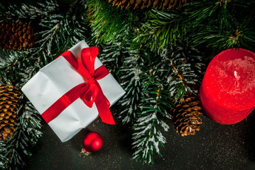 Christmas background for greeting card. Christmas tree branches with snow effect with festive red ribbon, pine cones, gift boxes and candles, on dark blue background, copy space top view