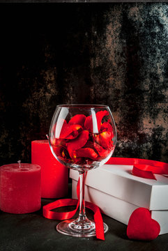 Valentine's Day Concept, White Wrapped Gift Box With Red Ribbon, Rose Flower Petals In Wine Glass, With Red Candle, On Dark Stone Background, Copy Space