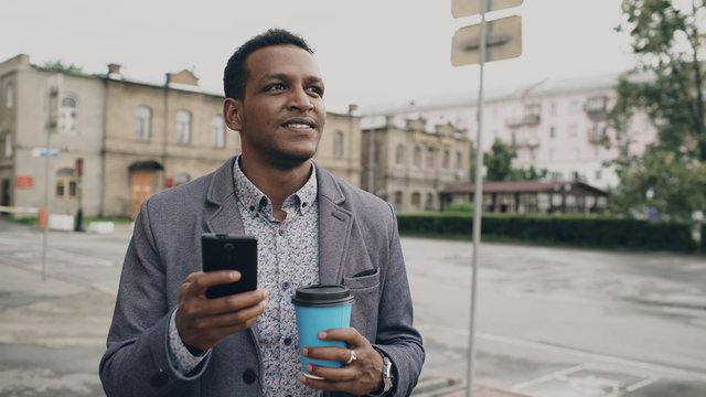 young happy businessman using smartphone and walking with cup of coffee outdoors