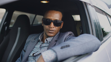Portrait of serious businessman in sunglasses sitting inside car and looking into camera outdoors