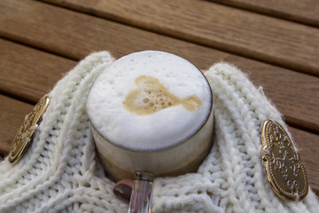 Girl hands in white mittens hold a cup of coffee with white foam and a heart on the background of a wooden table