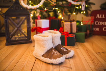 pair of warm home shoes made of wool stands near the Christmas tree with gifts at home on the wooden floor