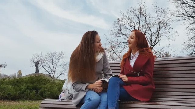 Female teenagers discussing a book sitting on the bench in an autumn city park