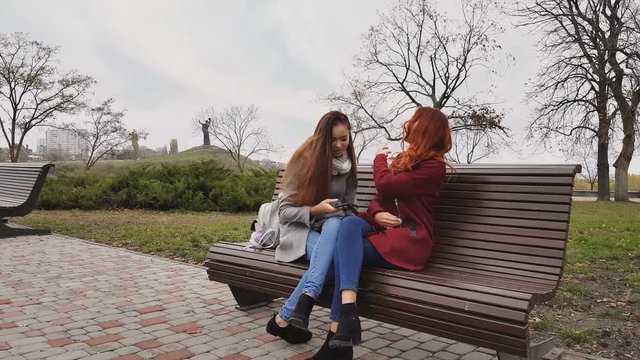 Female teenagers listening to music on smartphone sitting on the bench in an autumn city park
