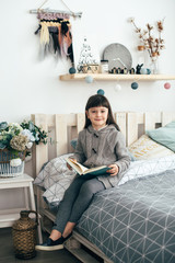 A little girl reads a book while sitting on the bed.
