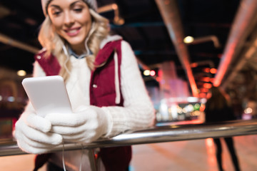 Fototapeta premium close-up view of smiling young woman in earphones using smartphone while standing on rink