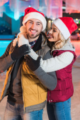 beautiful happy young couple in santa hats hugging on rink