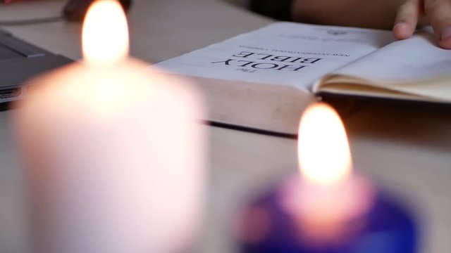 Man reading and studying Holy Bible lying on the table at home with blurry candles burning