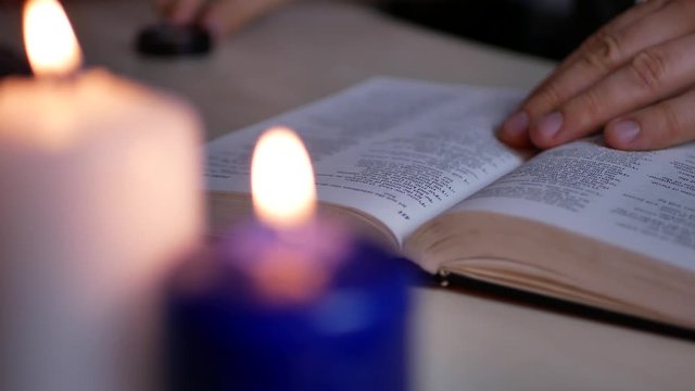 A man with magnifying ring reading and studying Holy Bible lying on the table with blurry candles burning