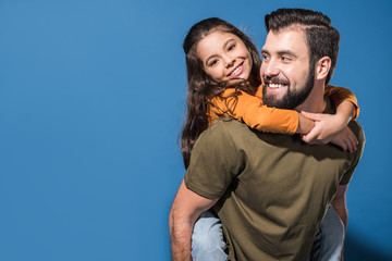 father giving piggyback to smiling daughter on blue