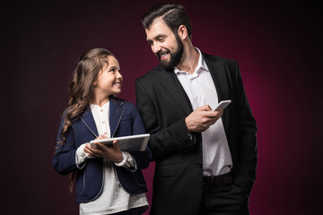 smiling father and daughter with tablet and smartphone looking at each other on burgundy