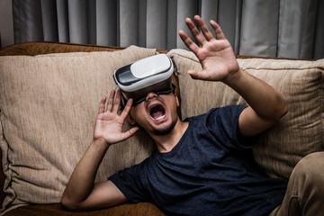 Asian man wearing virtual reality glasses expressing emotion screaming on brown sofa, smartphone using with VR glasses