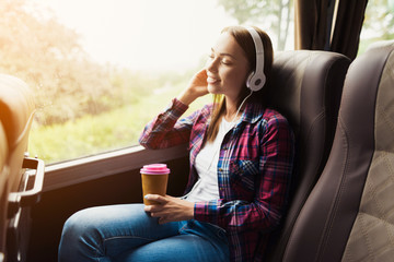 The woman on the passenger seat of the bus listens to music and drinks coffee.