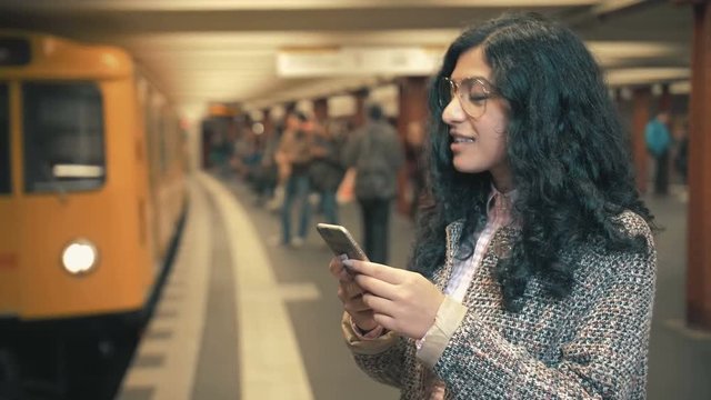 The girl of the mixed race awaits the train on the platform of the subway and touches the screen of the smartphone