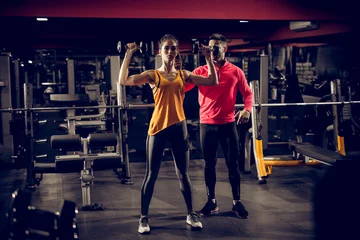 Fotobehang Strong motivated attractive muscular fitness girl standing with dumbbells above and doing a shoulder exercise while her handsome personal trainer standing behind and giving instructions in the gym. © dusanpetkovic1