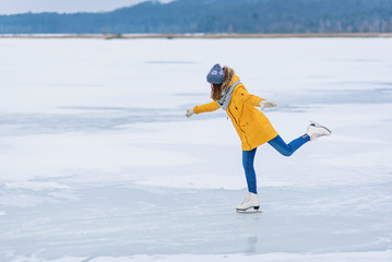 Young beautiful girl in yellow jacket is skating at winter on a frozen lake. Girl performs figure...