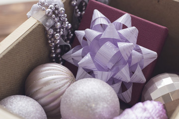 New Year decorations in a wooden box. Lilac shiny Christmas-tree balls, silver beads and a burgundy gift box with a lilac bow in the box. Close up